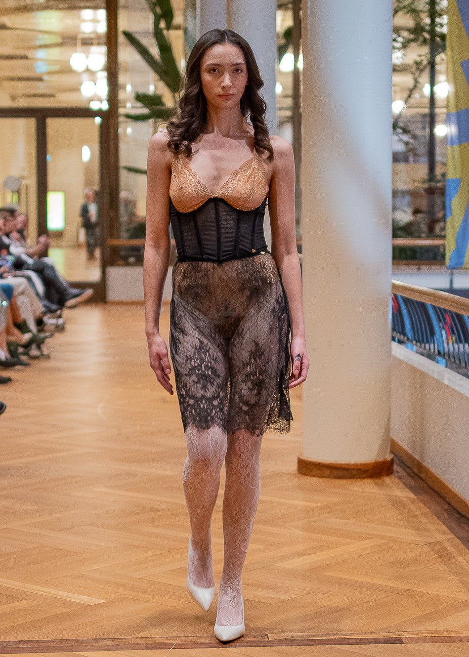 Model walking on a runway wearing a black and gold outfit with sheer elements.